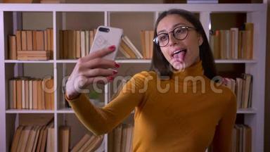 漂亮的深色头发的女老师<strong>戴</strong>着眼镜，在智能<strong>手机</strong>上做滑稽的<strong>自拍</strong>，在图书馆里欢快地露出舌头。